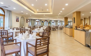 Un comedor espacioso con varias mesas dispuestas para el servicio, un mostrador de buffet con diferentes opciones de comida y grandes ventanales que dejan entrar luz natural.