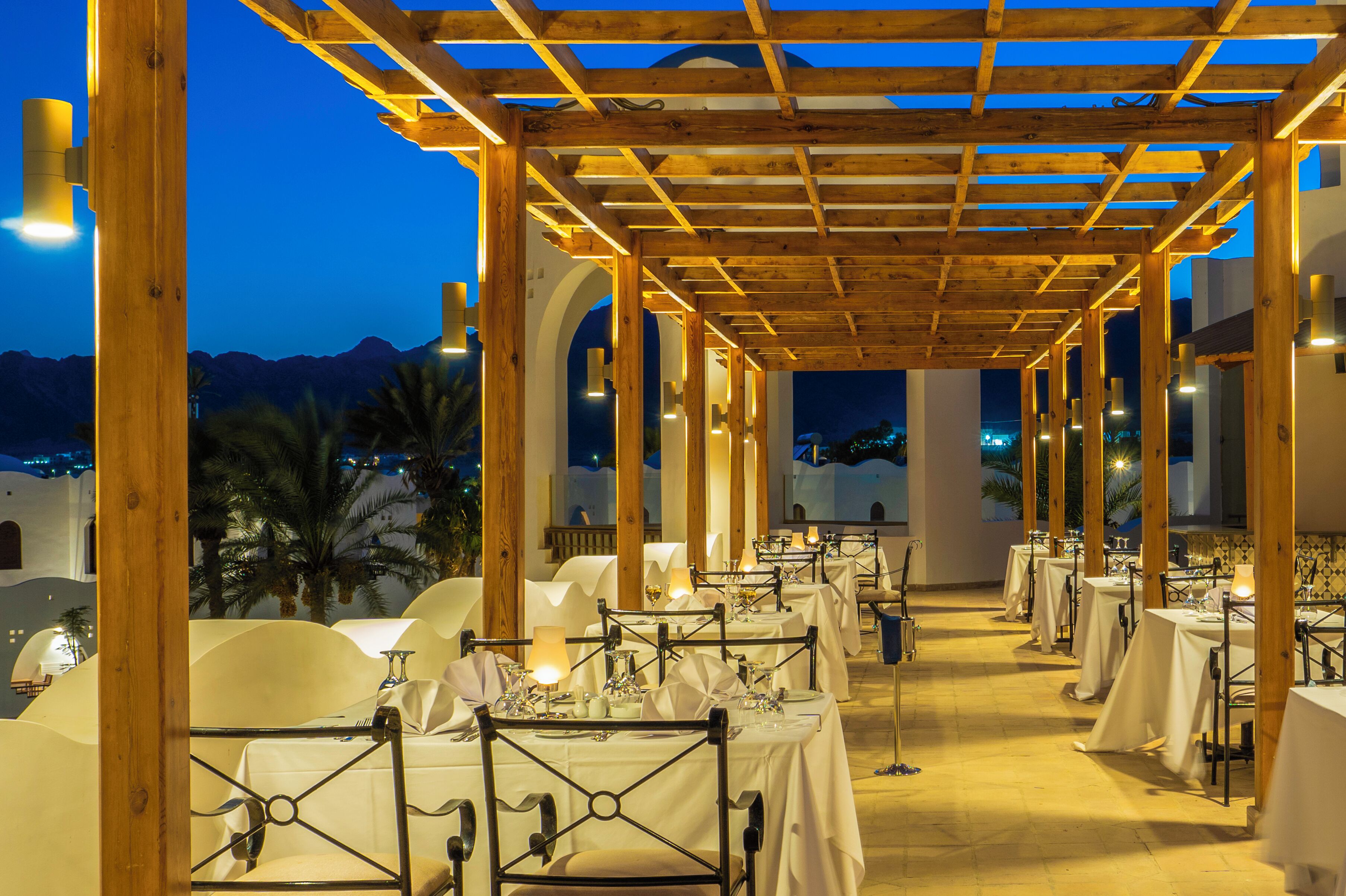 Un espacio de comedor al aire libre con mesas dispuestas para una comida, una pérgola de madera encima y vistas a las montañas al fondo.