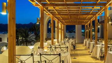 Un espacio de comedor al aire libre con mesas dispuestas para una comida, una pérgola de madera encima y vistas a las montañas al fondo.