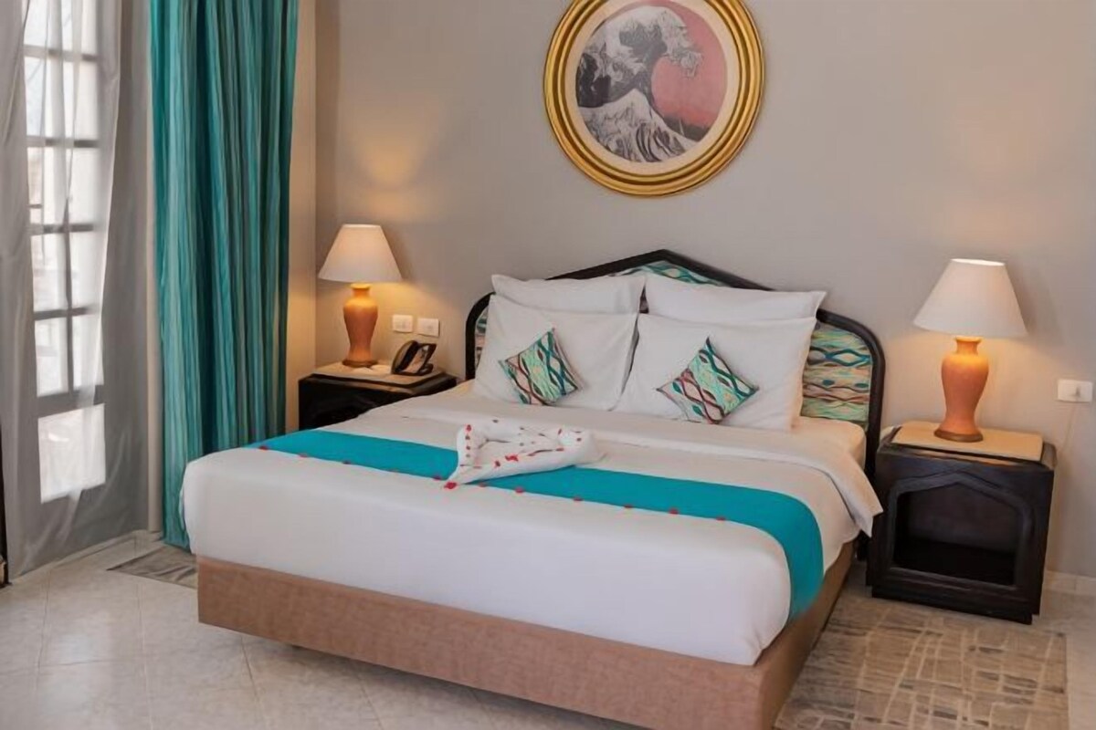 Una cama bien hecha con una colcha blanca y verde azulada, dos lámparas de noche y una obra de arte enmarcada sobre la cama.