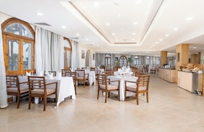 Un comedor amplio con varias mesas y sillas, ventanas grandes con cortinas y una estación de bufet.