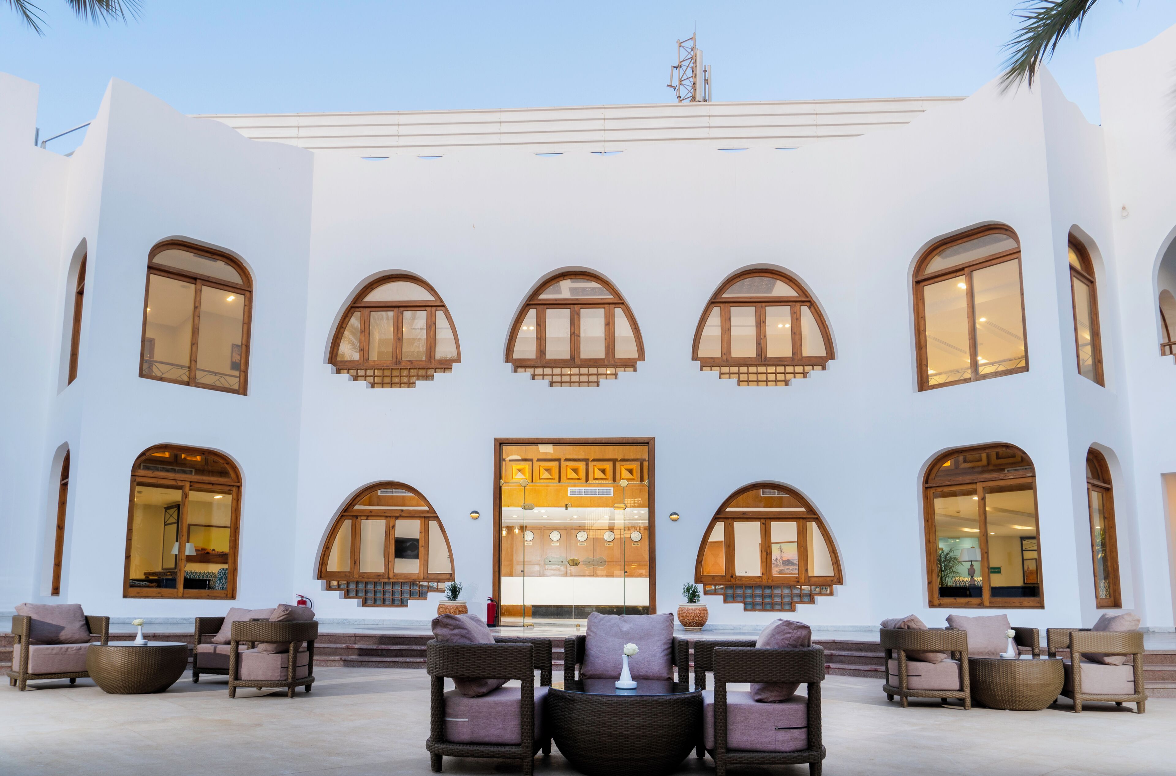 A building with multiple arched windows and a central door, outdoor seating area with wicker furniture, and a clear sky.