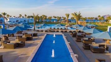 Uma área de piscina em resort, com várias espreguiçadeiras e guarda-sóis, rodeada por palmeiras e vista clara do oceano.