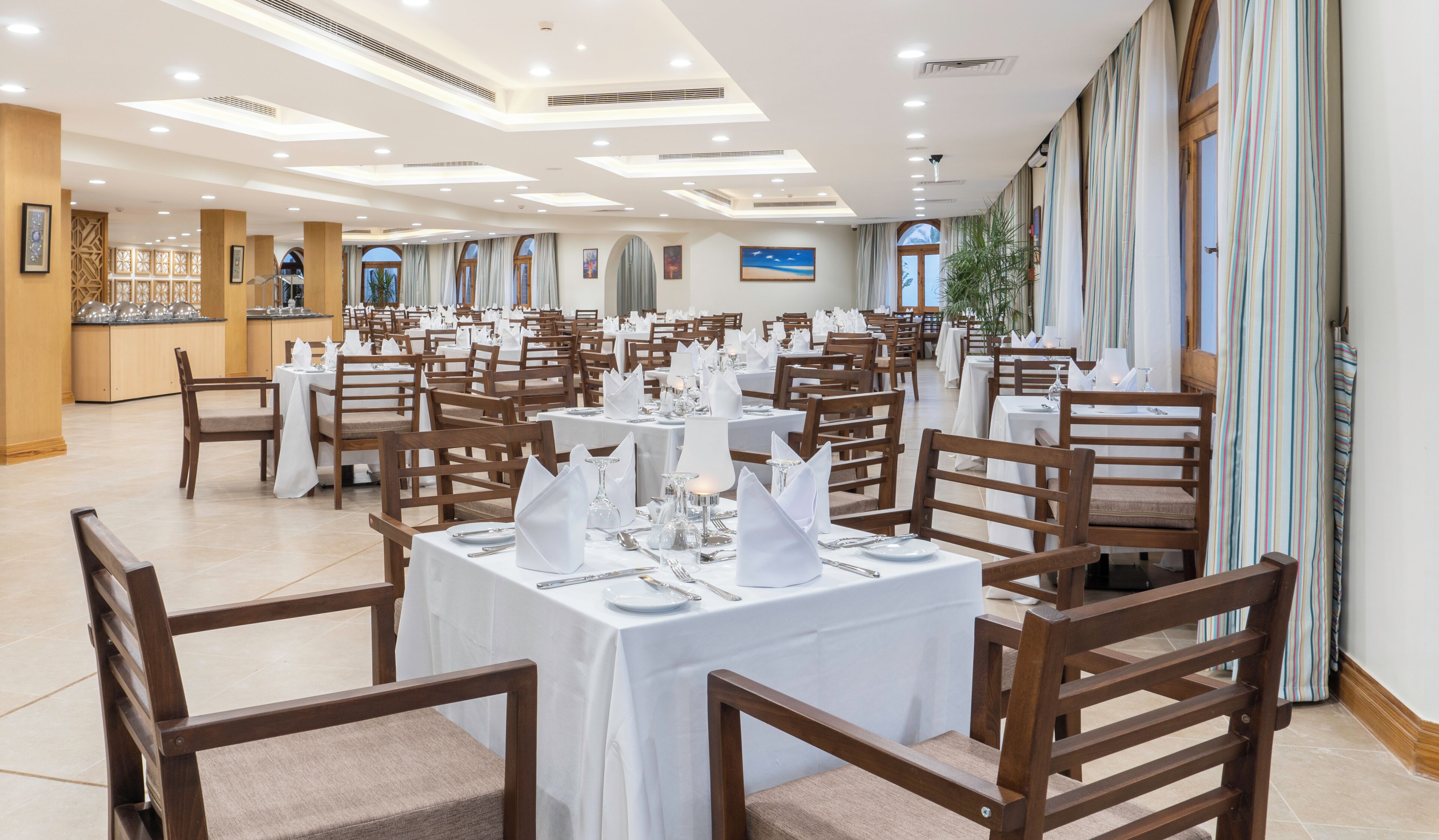 A spacious dining area with multiple tables set for service, a kitchen in the background, and a large window providing natural light.