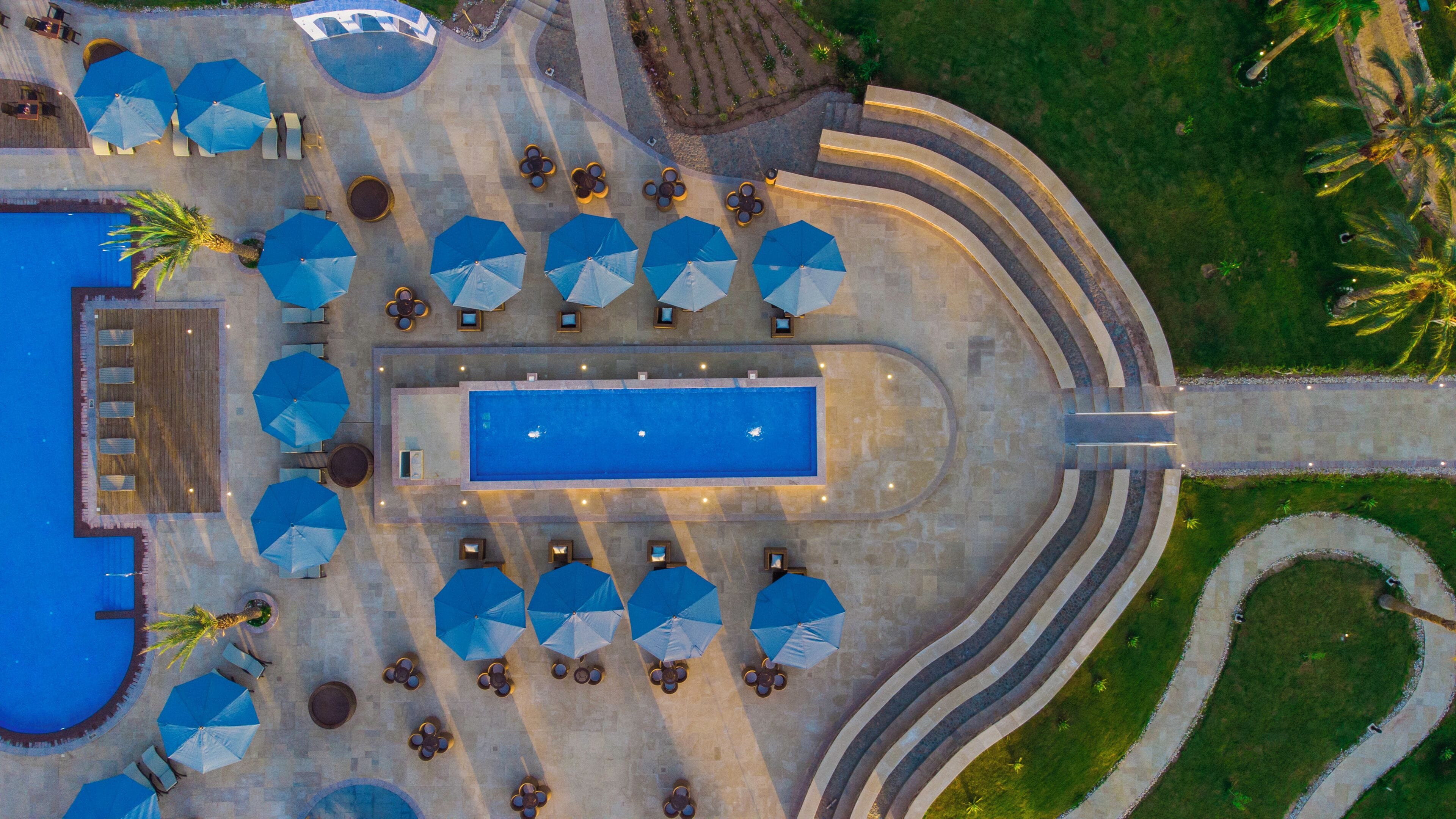 An aerial view of a resort with a central swimming pool, sun loungers, and umbrellas.