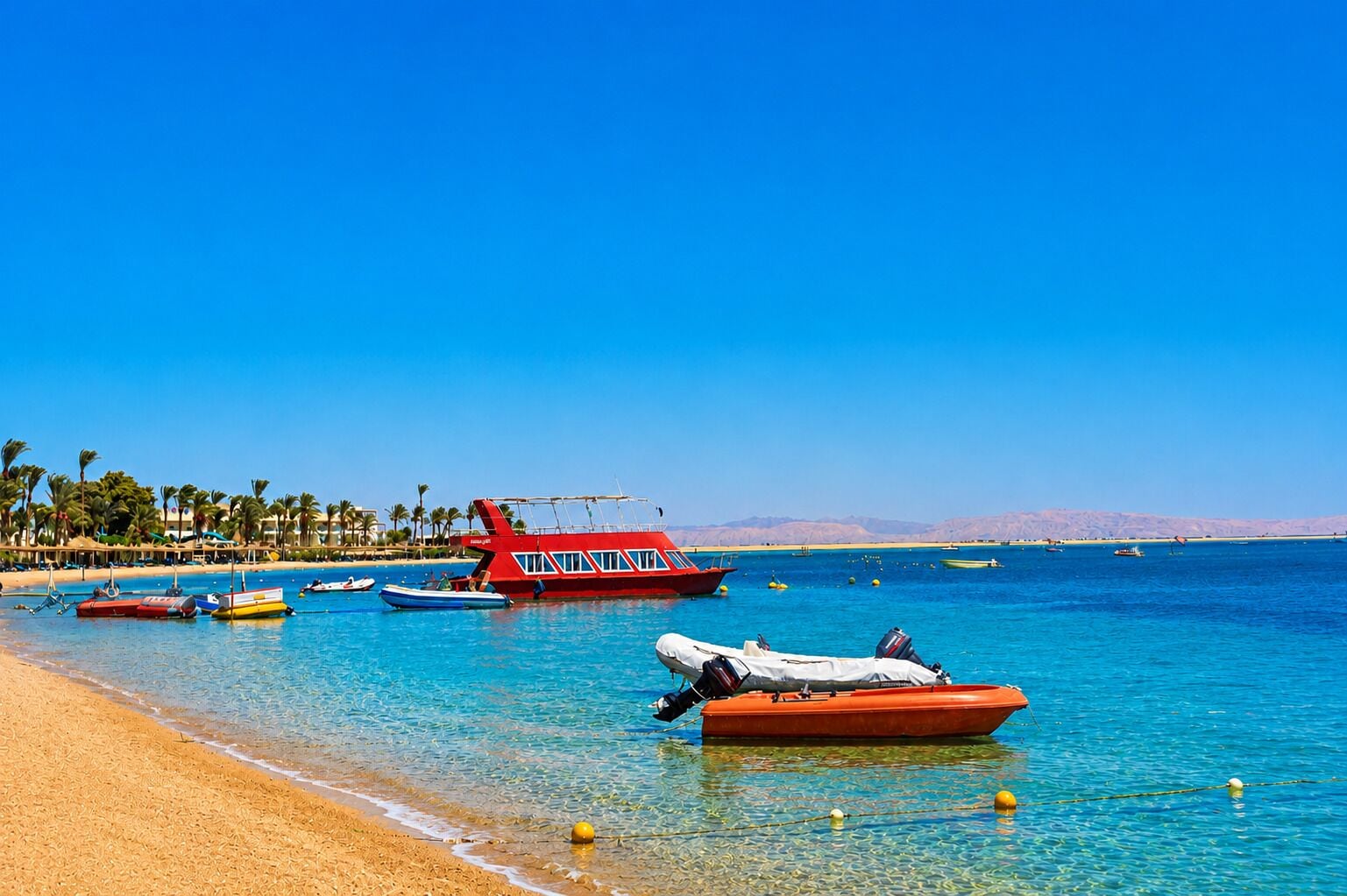 A beach with clear blue water, several boats, and a sandy shore with palm trees.