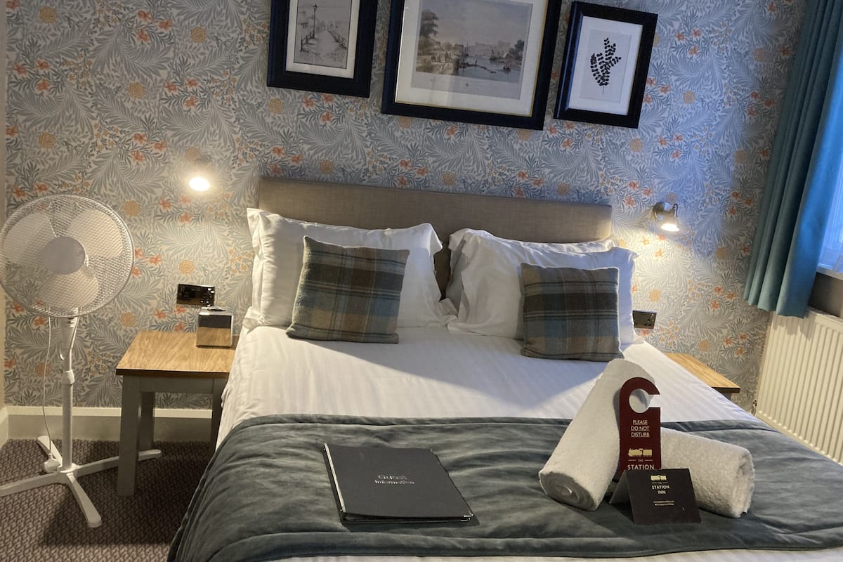 A hotel room with a bed, pillows, a bedside table, a fan, and framed pictures on the wall.
