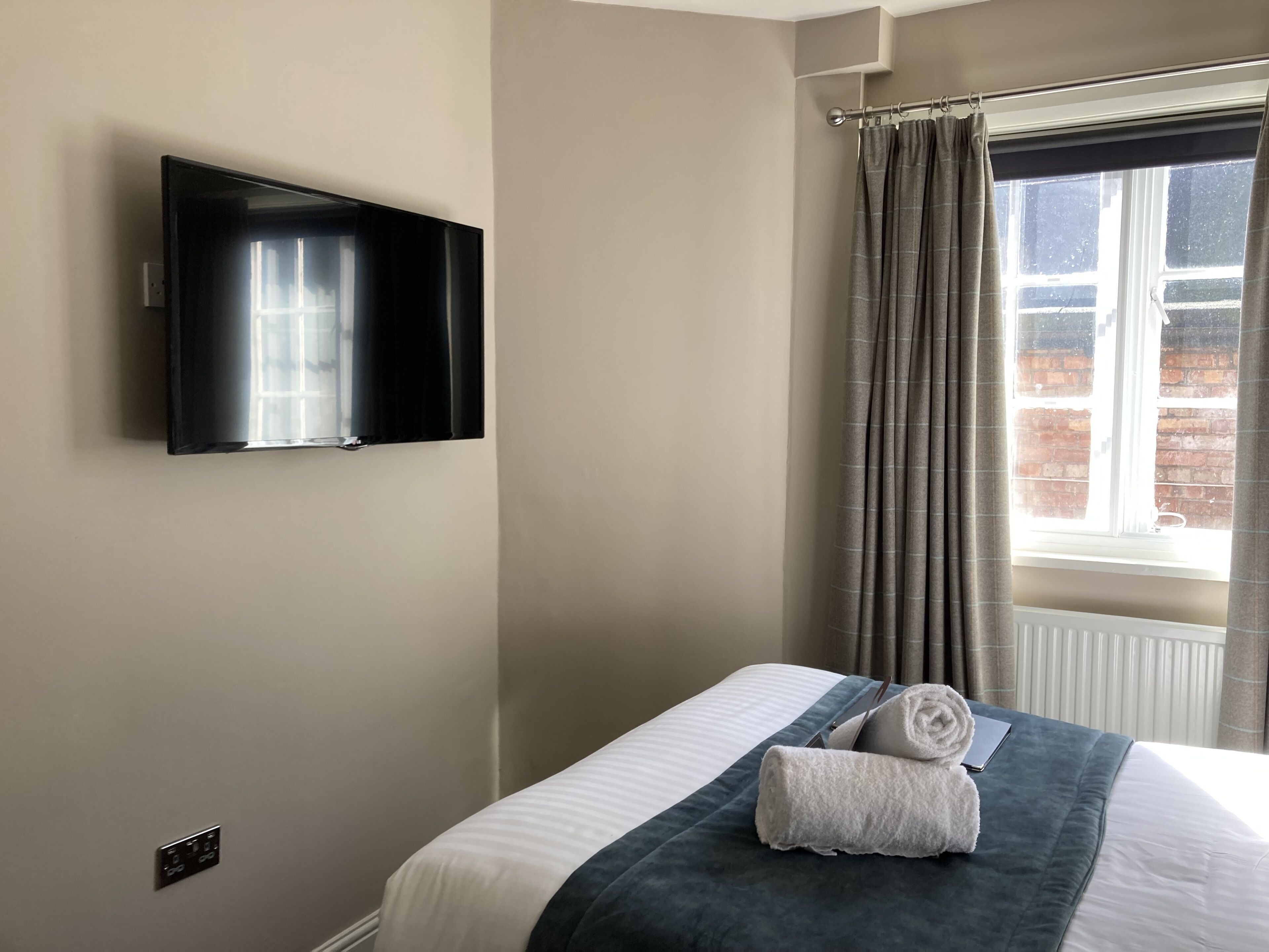 A hotel room with a bed, a flat-screen TV mounted on the wall, a window with curtains, and a view of the outside.