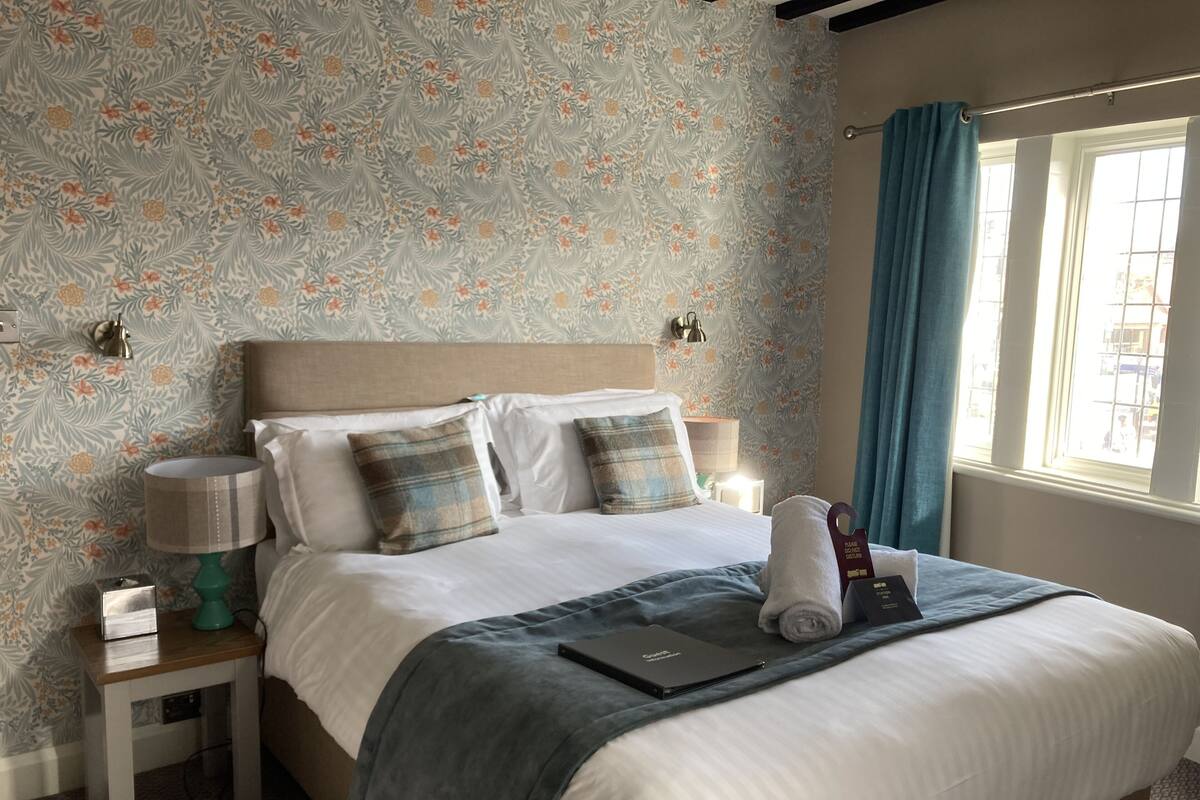 A neatly made bed with a blue blanket, two pillows, a bedside lamp, and a small table with a book and a tissue box.