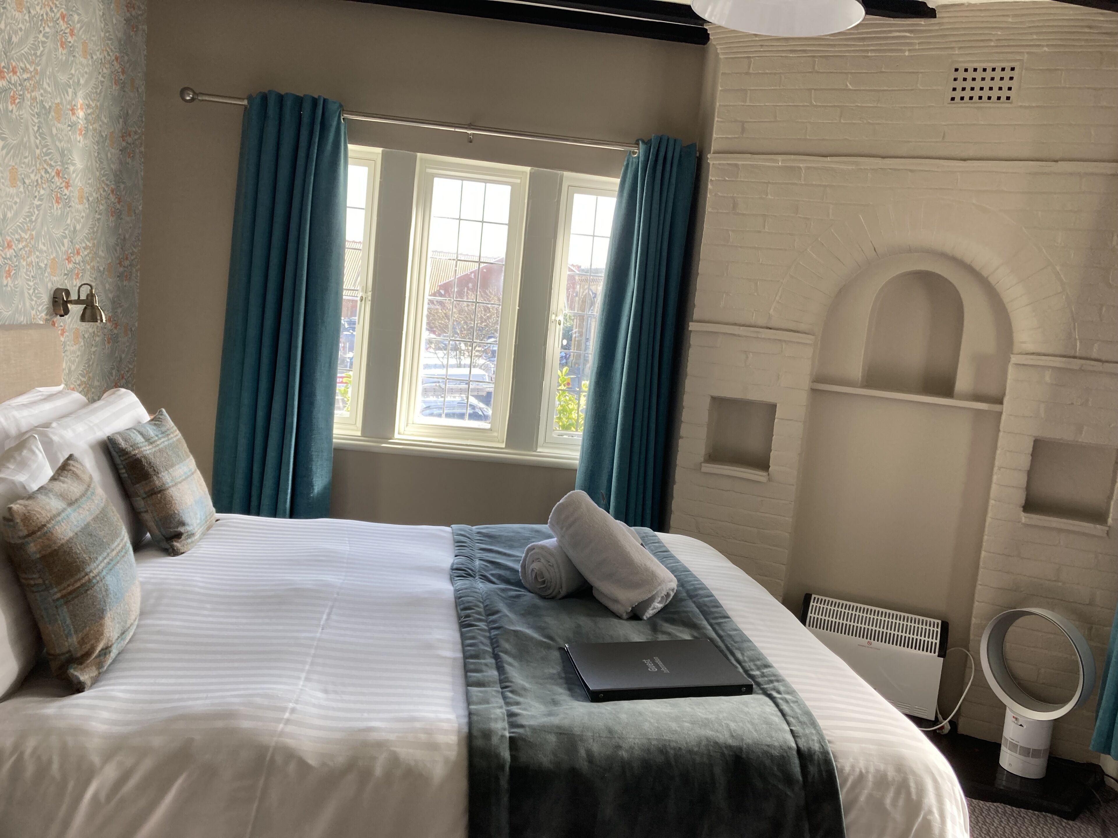 A neatly made bed with a laptop, blue curtains, and a white brick wall with a fireplace.