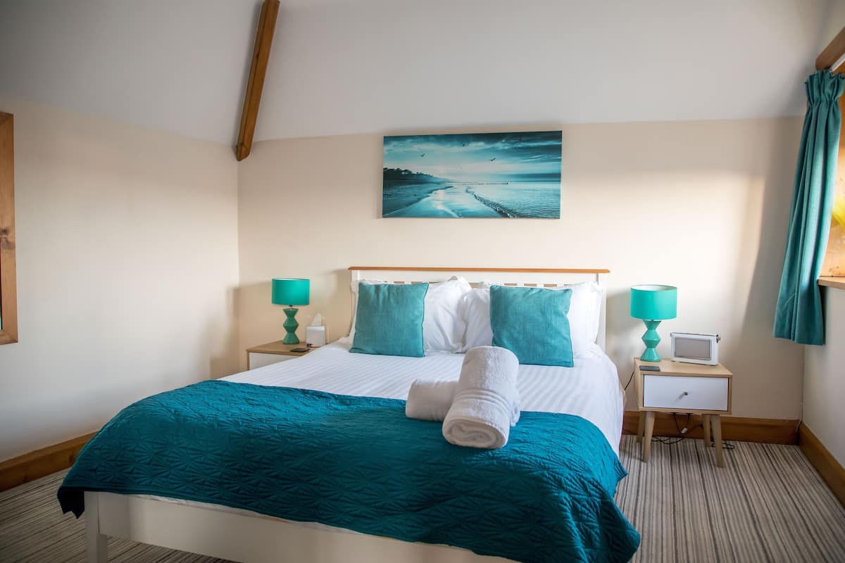 A neatly made bed with a teal bedspread, two matching pillows, and a rolled towel. There is a small bedside table with a lamp and a television. A painting of a coastal scene hangs above the bed.
