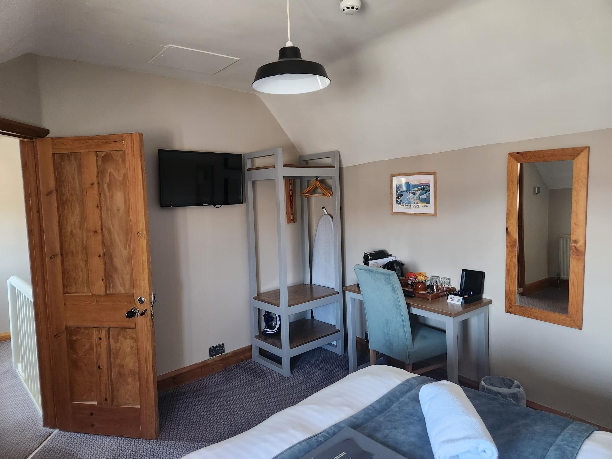 A bedroom with a bed, a desk, a chair, a TV, a mirror, and a door.