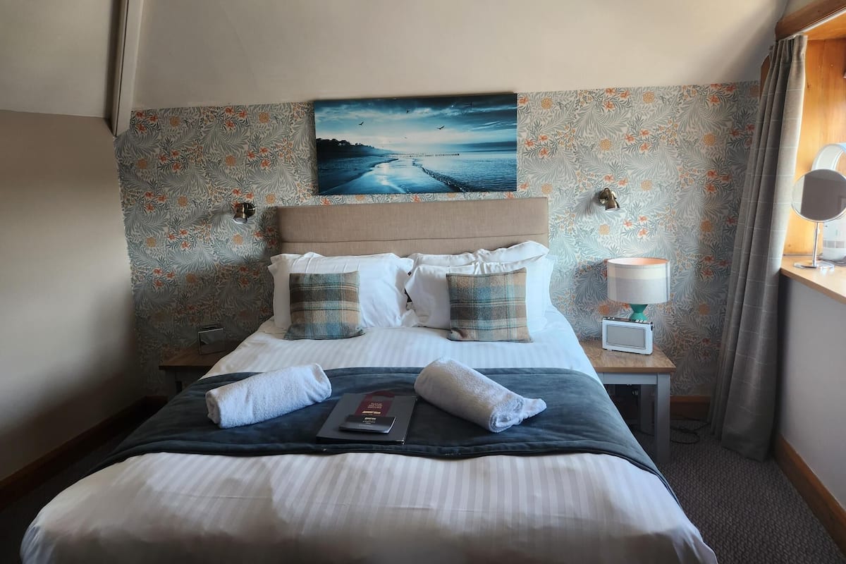A neatly made bed with white linens and two plaid pillows, a bedside table with a lamp, and a painting of a seascape on the wall.