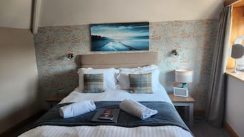Una cama bien hecha con sábanas blancas y dos almohadas de cuadros, una mesita de noche con una lámpara, y un cuadro de un paisaje marino en la pared.