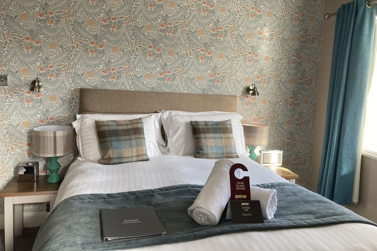 A neatly made bed with a floral wallpaper, a bedside lamp, a folded towel, a guest information booklet, and a 'Please Do Not Disturb' sign.
