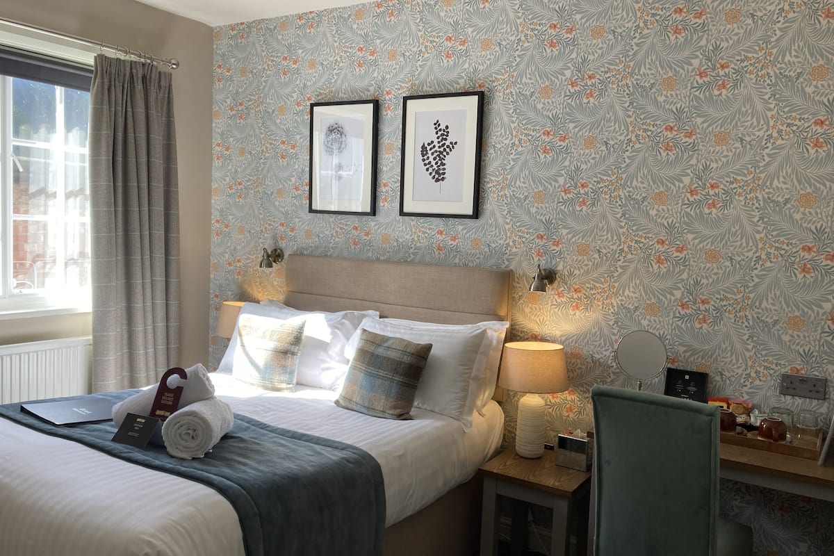 A hotel room with a bed, a desk, a chair, a window with curtains, and two framed pictures on the wall.