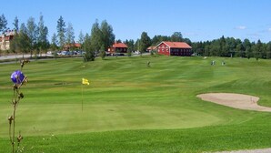 Ein Golfplatz mit Fahne, Sandbunkern und roten Gebäuden im Hintergrund.