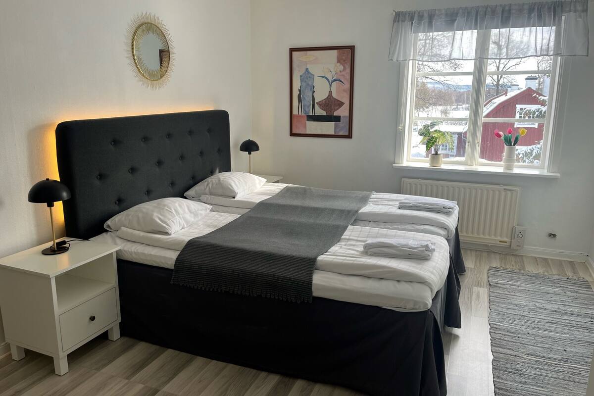 A bedroom with a large bed, two bedside tables, a floor lamp, a window with a view, and a wall-mounted mirror.