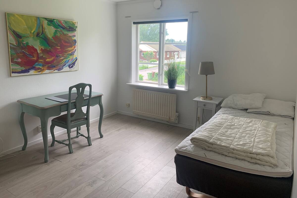 A bedroom with a desk, chair, bed, window, radiator, and a painting.