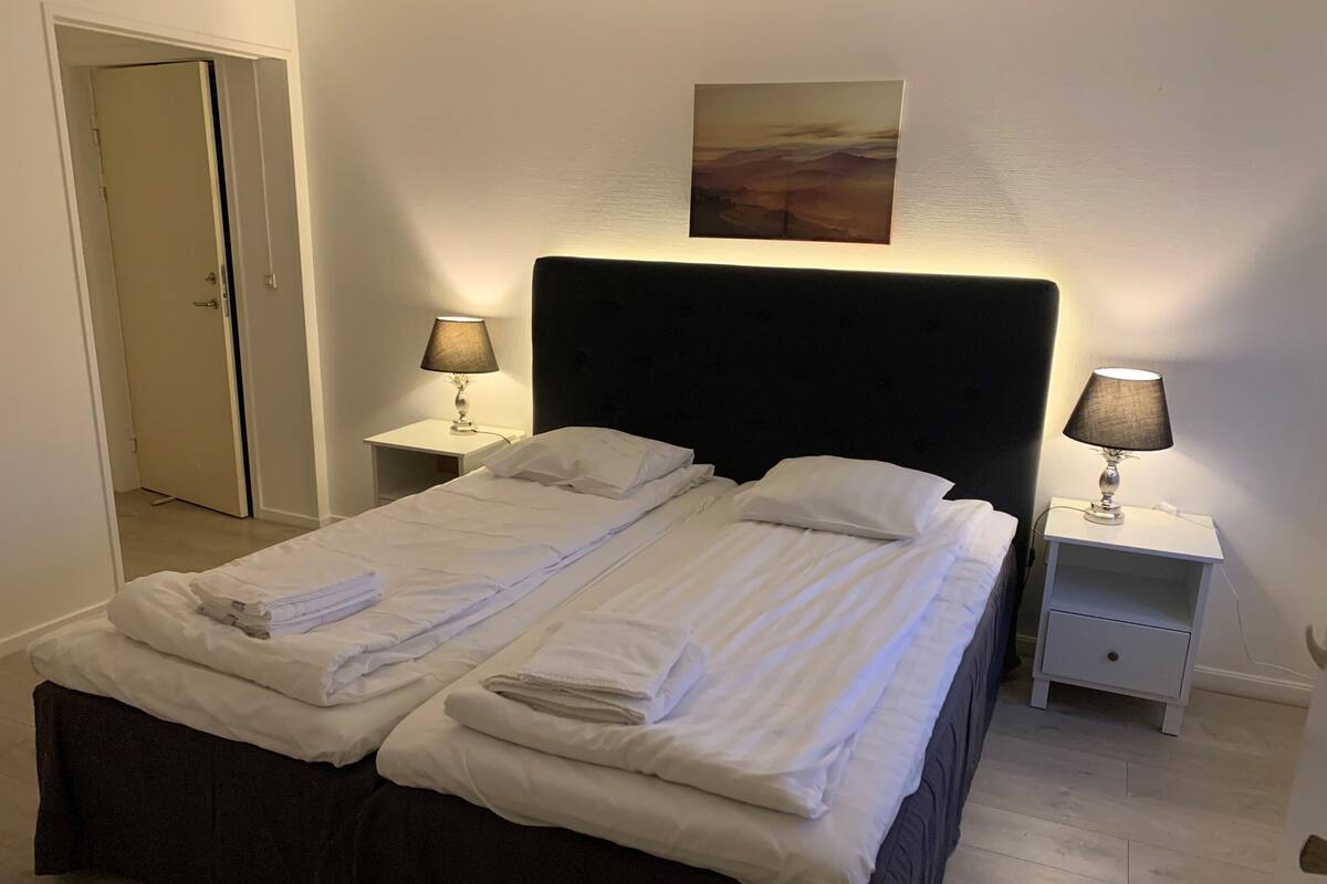 A double bed with white linens, two bedside tables with lamps, a painting on the wall, and a door in the background.