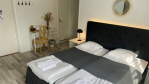 Ein ordentlich bezogenes Bett mit weißen Leinentüchern und einer grauen Decke, ein Nachttisch mit einer Lampe, ein Stuhl und ein kleiner Tisch mit einer Blumenvase.
