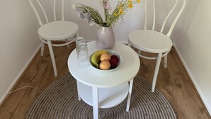 Ein kleiner runder Tisch mit einer weißen Vase mit Blumen und einer Obstschale. Zwei weiße Stühle befinden sich auf beiden Seiten des Tisches.