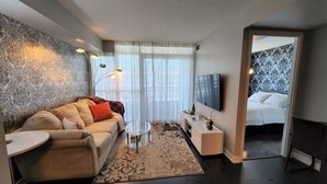 Ein modernes Wohnzimmer mit Sofa, Couchtisch, Fernseher und Blick ins Schlafzimmer.