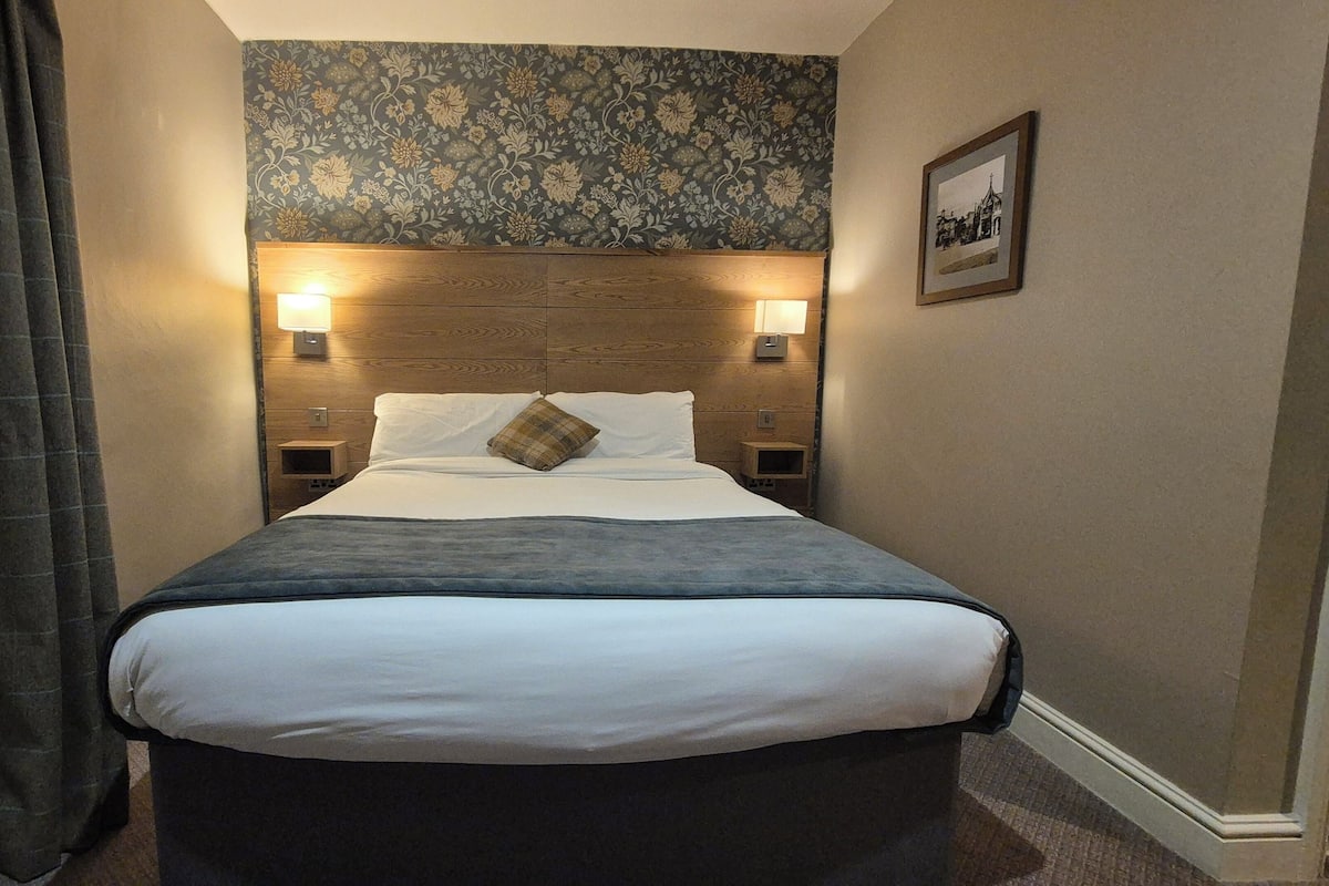 Ein Hotelzimmer mit einem großen Bett, zwei Nachttischlampen, einem mit floralem Muster versehenen Kopfteil und einem gerahmten Bild an der Wand.
