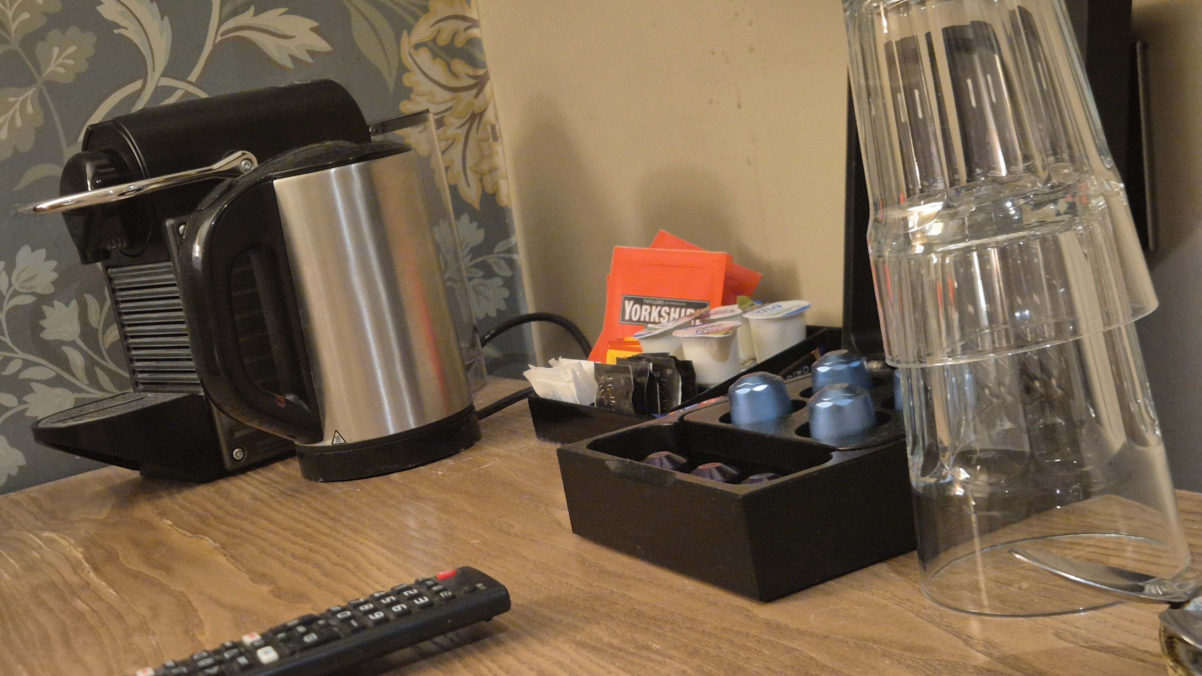 Una encimera de cocina con una cafetera, un hervidor, una jarra de vidrio, un soporte para hervidor y un control remoto.