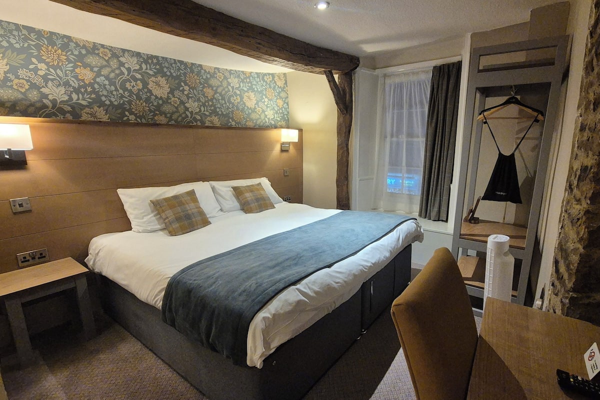 Ein Hotelzimmer mit einem großen Bett, einem Holzkopfende, einem Nachttisch, einem Schreibtisch mit Stuhl und einem fenster mit Vorhängen.