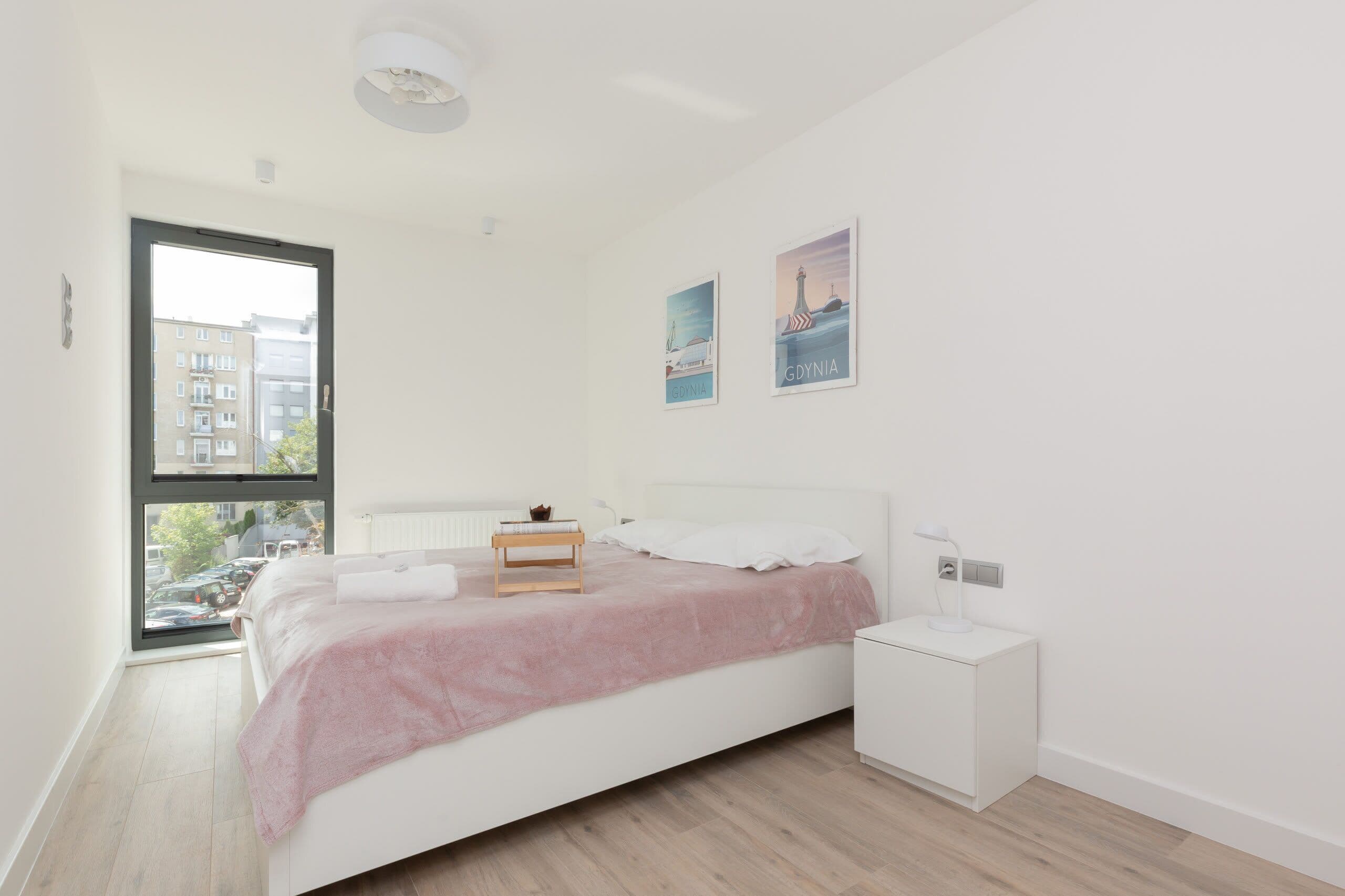 A modern bedroom with a large window, a bed with a pink blanket, a bedside table with a lamp, and two wall art pieces.