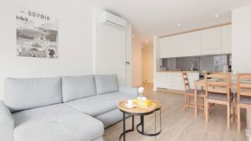 A modern living room with a grey sofa, a small round table with a vase and a cup, and a kitchen area with wooden cabinets and a dining table.