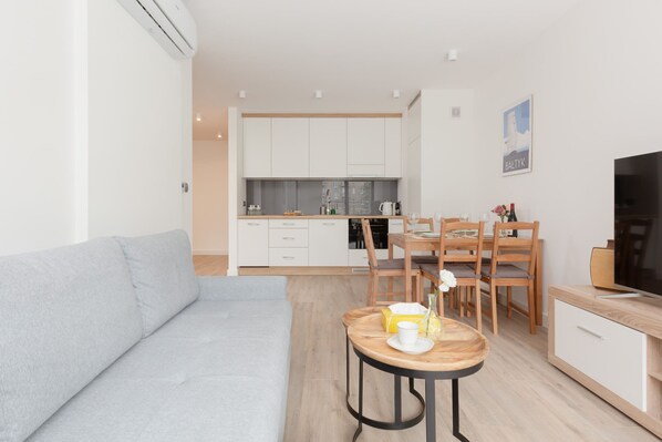 A modern living room with a grey sofa, a wooden dining table with chairs, a kitchen area with white cabinets, and a TV on a wooden stand.