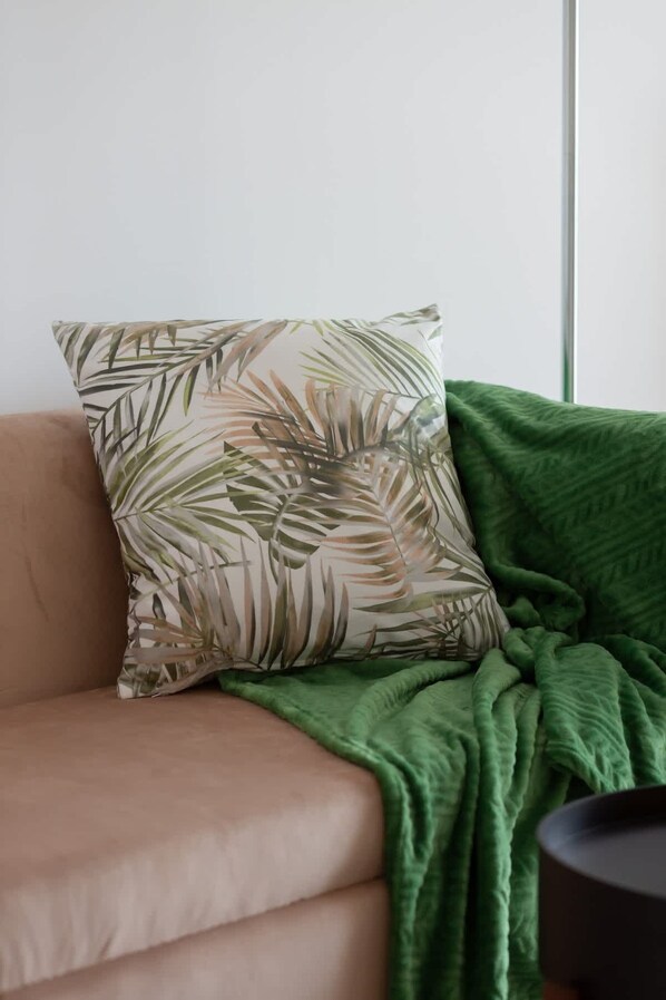 Un sofá color beige con un cojín que presenta un diseño de hojas tropicales. Una manta verde está colocada sobre el sofá.
