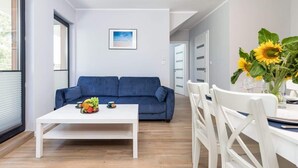 A modern living room with a blue sofa, a white coffee table with fruit, and a dining area with white chairs and a vase of sunflowers. - Modern Apartment | 550 m to the Beach | Parking | Spa (Międzywodzie)