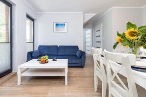 Un salon moderne avec un canapé bleu, une table basse blanche ornée de fruits et un coin repas avec des chaises blanches et un vase rempli de tournesols.
