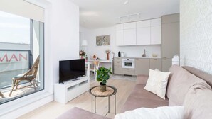 Ein modernes Wohnzimmer mit einer Couch, einem Fernseher und einem kleinen Tisch mit einer Pflanze. Im Hintergrund ist eine Küche mit Schränken und einem Essbereich zu sehen.