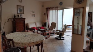 A living room with a dining table, chairs, a sofa, and a chair. There is a window with curtains, a ceiling fan, and a wall-mounted shelf with decorative items. - Spacious villa close to center and beaches. (Hyères)
