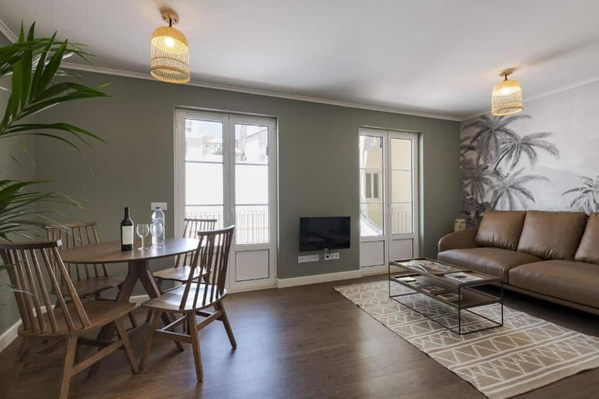 Un salon avec un canapé en cuir marron, une table à manger avec des chaises, un téléviseur fixé au mur et de grandes fenêtres.