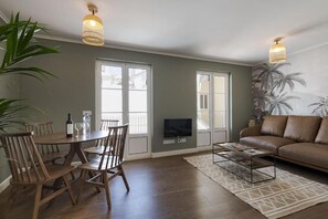 Een woonkamer met een bruine leren bank, een eettafel met stoelen, een aan de muur bevestigde televisie en grote ramen.