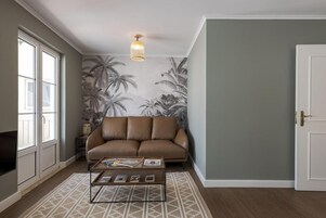 Una sala de estar con un sofá de cuero marrón, una mesa de centro con revistas, un tapete estampado y una pared con papel tapiz tropical.
