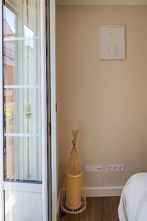 Een kamer met een grote schuifpui, een geweven rotan stoel, een kunstwerk in een lijst aan de muur en een bed met een witte dekbedovertrek.