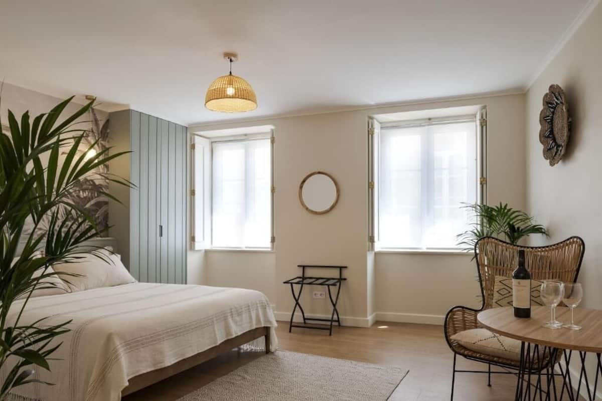 Une chambre à coucher avec un lit, une table avec des verres à vin, une chaise, un luminaire suspendu et un miroir rond accroché au mur.