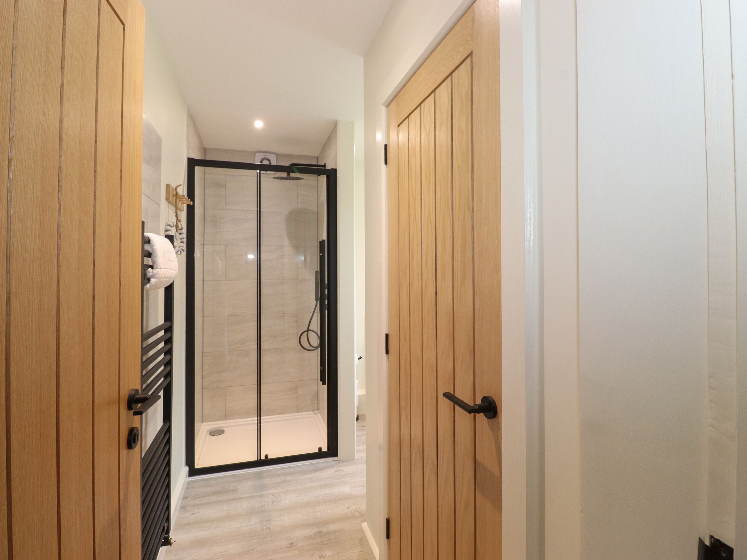 Un couloir mène à un espace de douche clos par des parois de verre, avec des portes en bois de chaque côté.