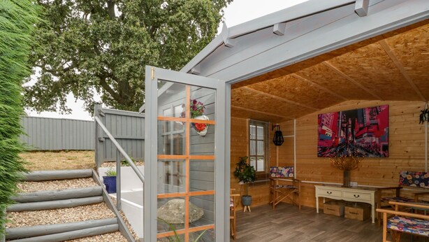 Una cabaña de madera con una escalera que lleva a una puerta de cristal, una zona de comedor con una mesa y sillas, y un cuadro llamativo en la pared.
