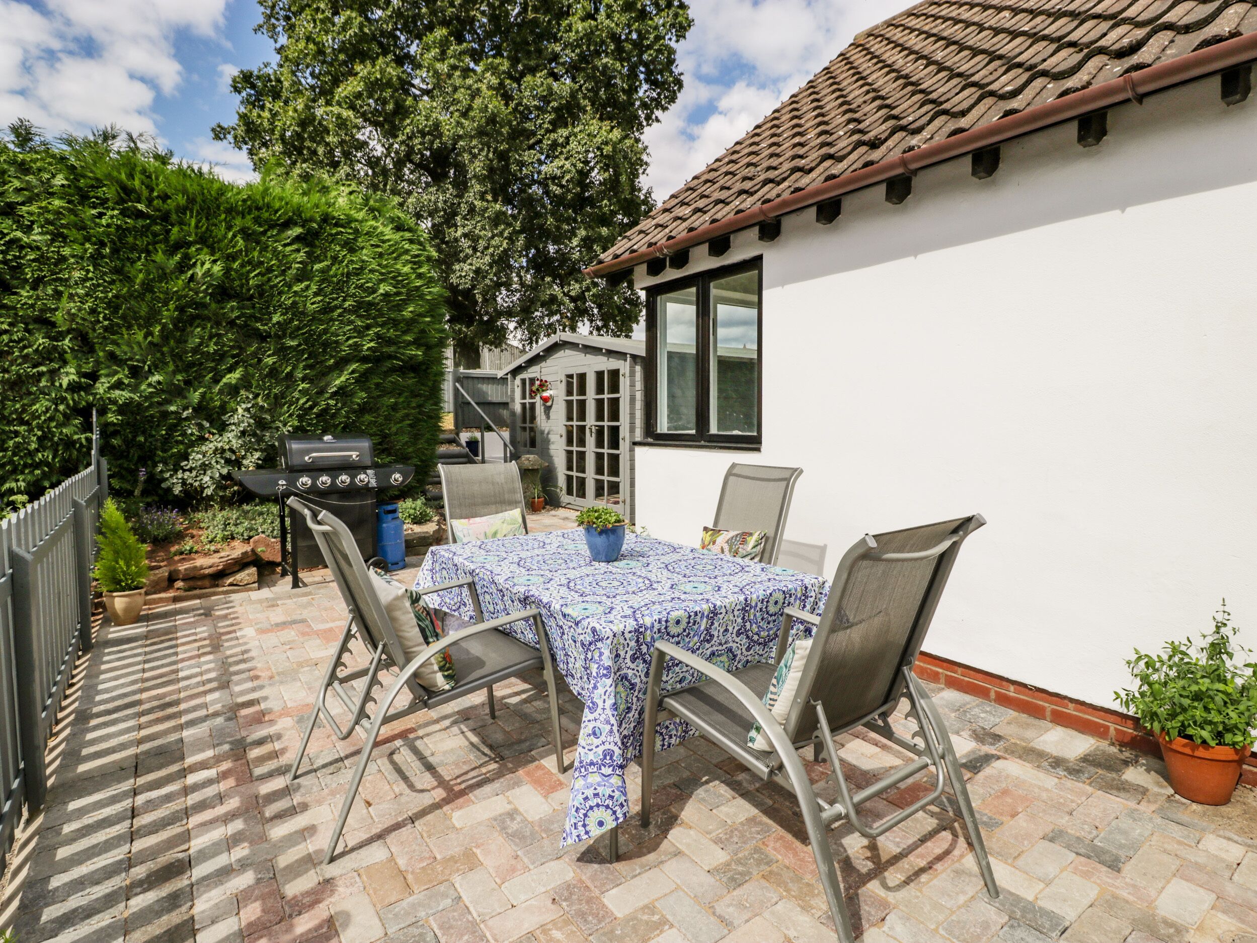 Une terrasse avec une table et des chaises, un barbecue et un espace jardin agrémenté d’arbres et de plantes.