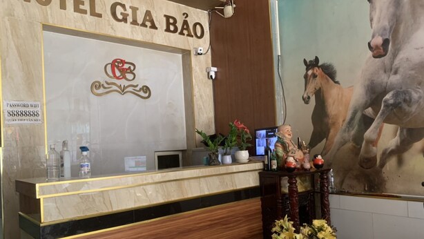 A hotel reception desk with a large sign reading "HOTEL GIÁ BÁO". There is a logo above the sign, a computer monitor on the desk, and a large horse painting on the wall to the right. - Gia Bao Hotel (Ho Chi Minh City)