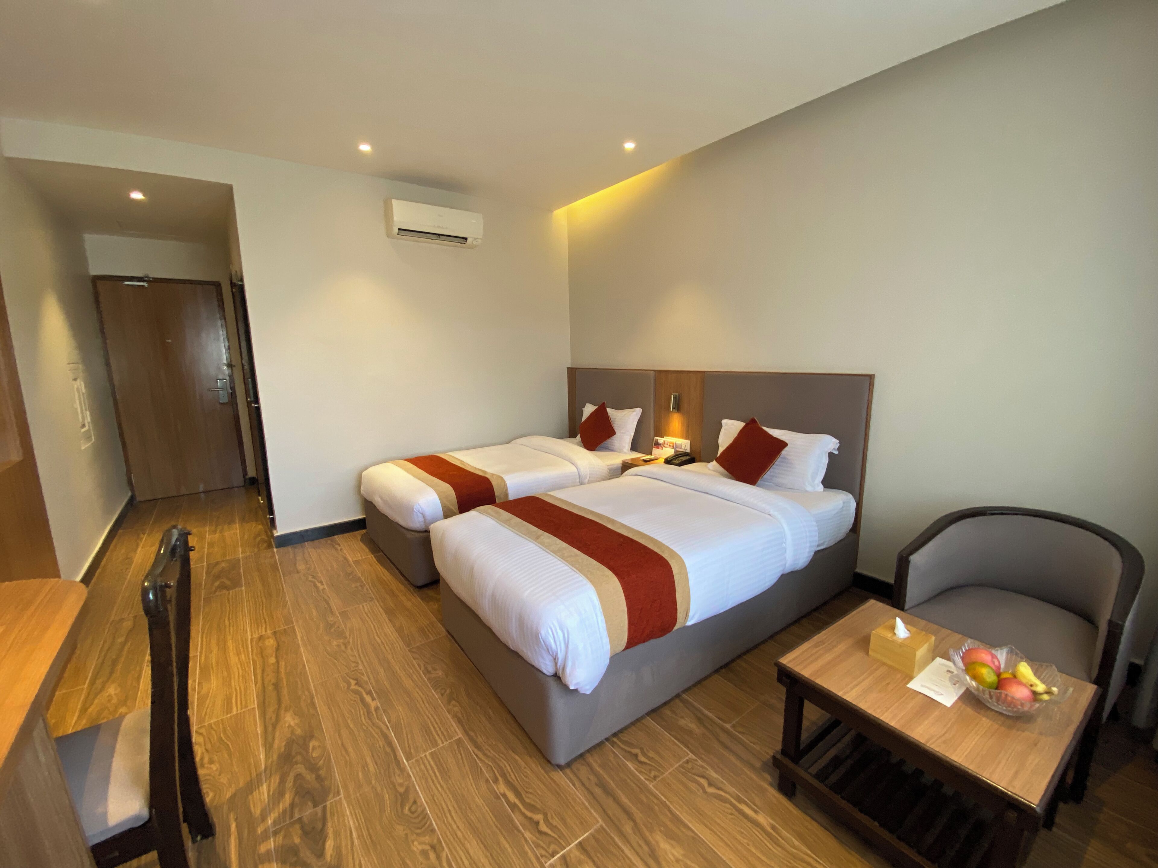 Quarto de hotel com duas camas, uma escrivaninha, uma cadeira, uma mesa com frutas e um aparelho de ar condicionado.