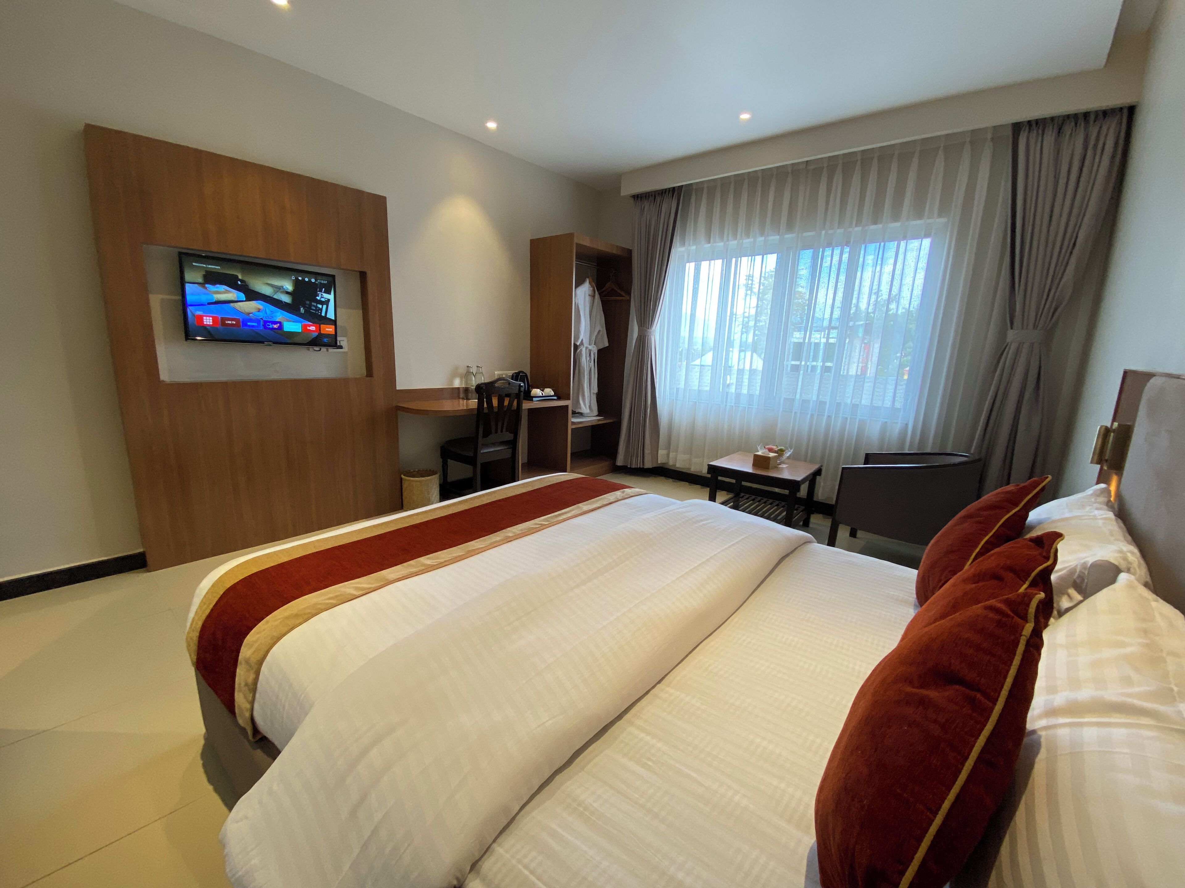 A hotel room with a large bed, a TV mounted on the wall, a desk with a chair, and a window with curtains.
