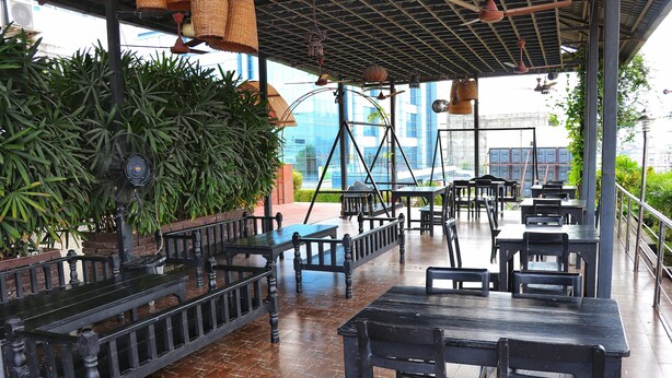 Um café ao ar livre com mesas e bancos de madeira, rodeado por vegetação e edifícios modernos ao fundo.